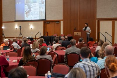 A group of people gather to listen to Paulette Granberry Russell deliver a keynote address. She is placed at a podium in the front of the room. 