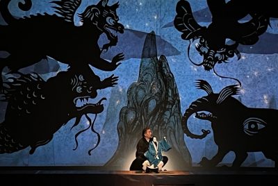 A Japanese man crouches on the floor of a stage holding a puppet while surrounded by animated drawings of dragons and other animals.