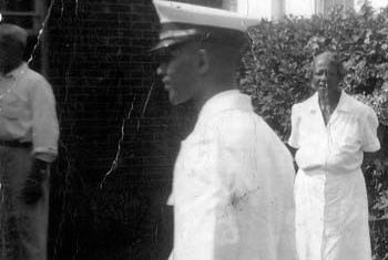 Essex Finney, a your Black cadet, is pictured in white dress uniform walking in front of Janie Hoge, anolder Black woman in a white dress. They are standing in the sun, outside in front of a brick building. 