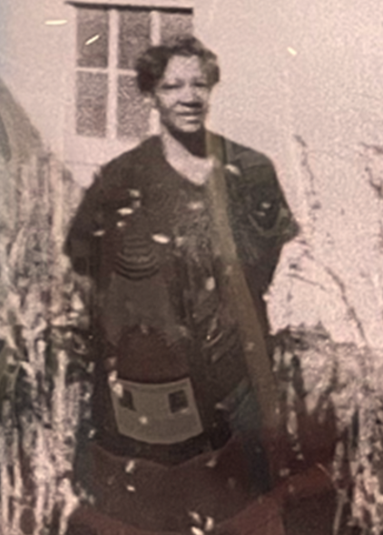 A Black woman with short hair stands in the sun wearing a dark, knee-length dress, her hands clasped behind her back. The photograph is black and white, grainy, clearly old. 